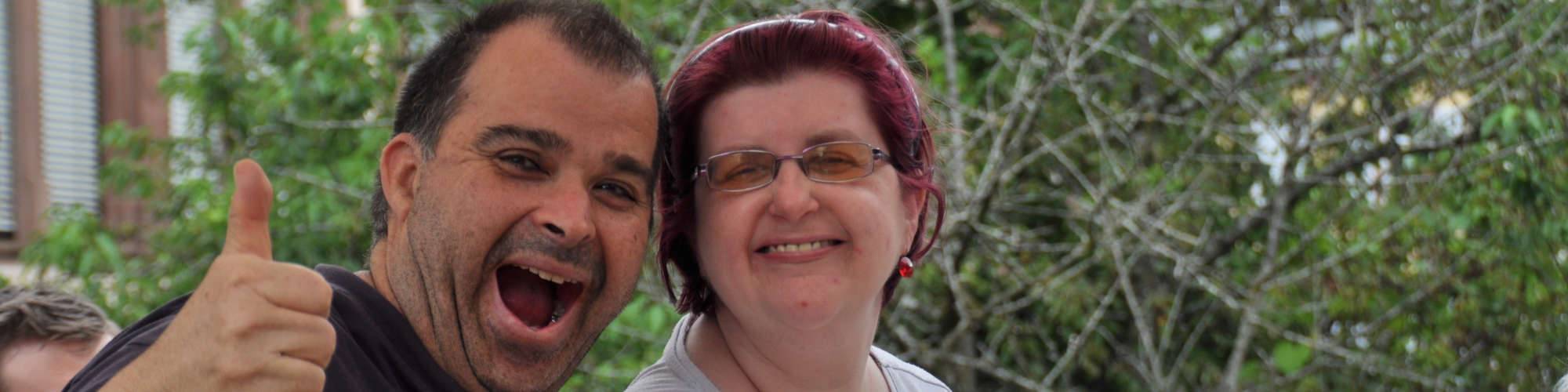 Ein Mann und eine Frau lächeln uns an, der Mann zeigt den Daumen nach oben, im Hintergrund ist das Grüne von einem Baum zu sehen