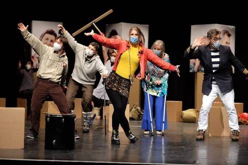 Gruppe maskierter Personen posiert auf Bühne, teils mit Requisiten, in lebhafter Szene.