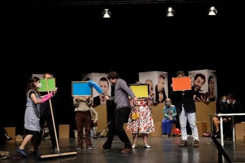 Personen auf Bühne halten bunte Tafeln, andere sitzen im Hintergrund, lebhafte Szene.